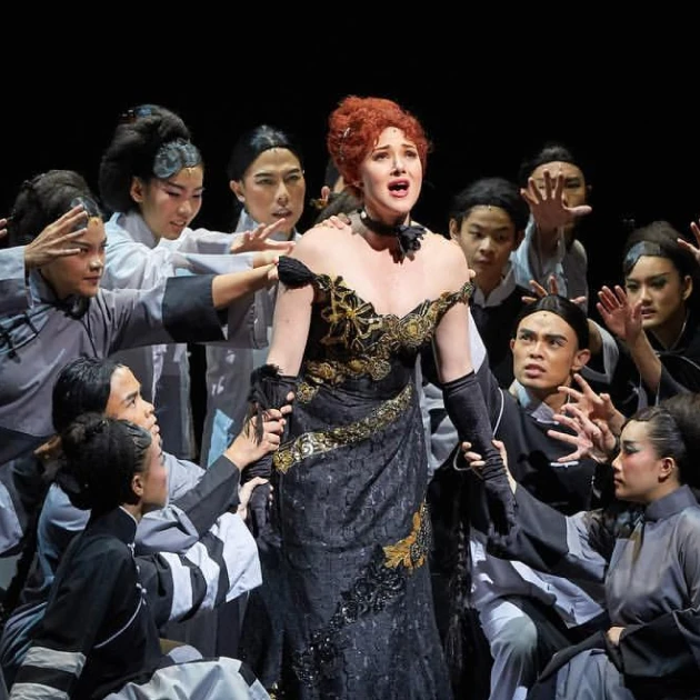 female singer wearing an extravagant black dress singing on stage surrounded by many performers