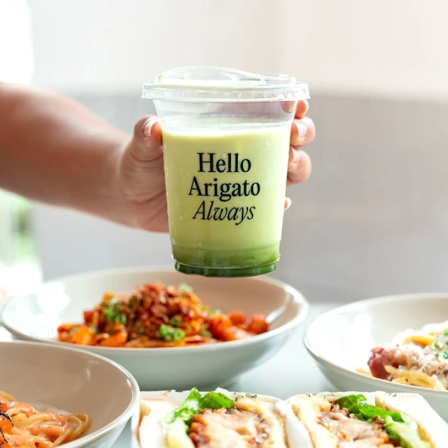 person holding a drink in a plastic cup with the words "hello arigato always" over a few plates of dishes
