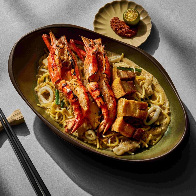 an aesthetic plate of hokkien mee with prawns and roast pork and sambal chilli on the side